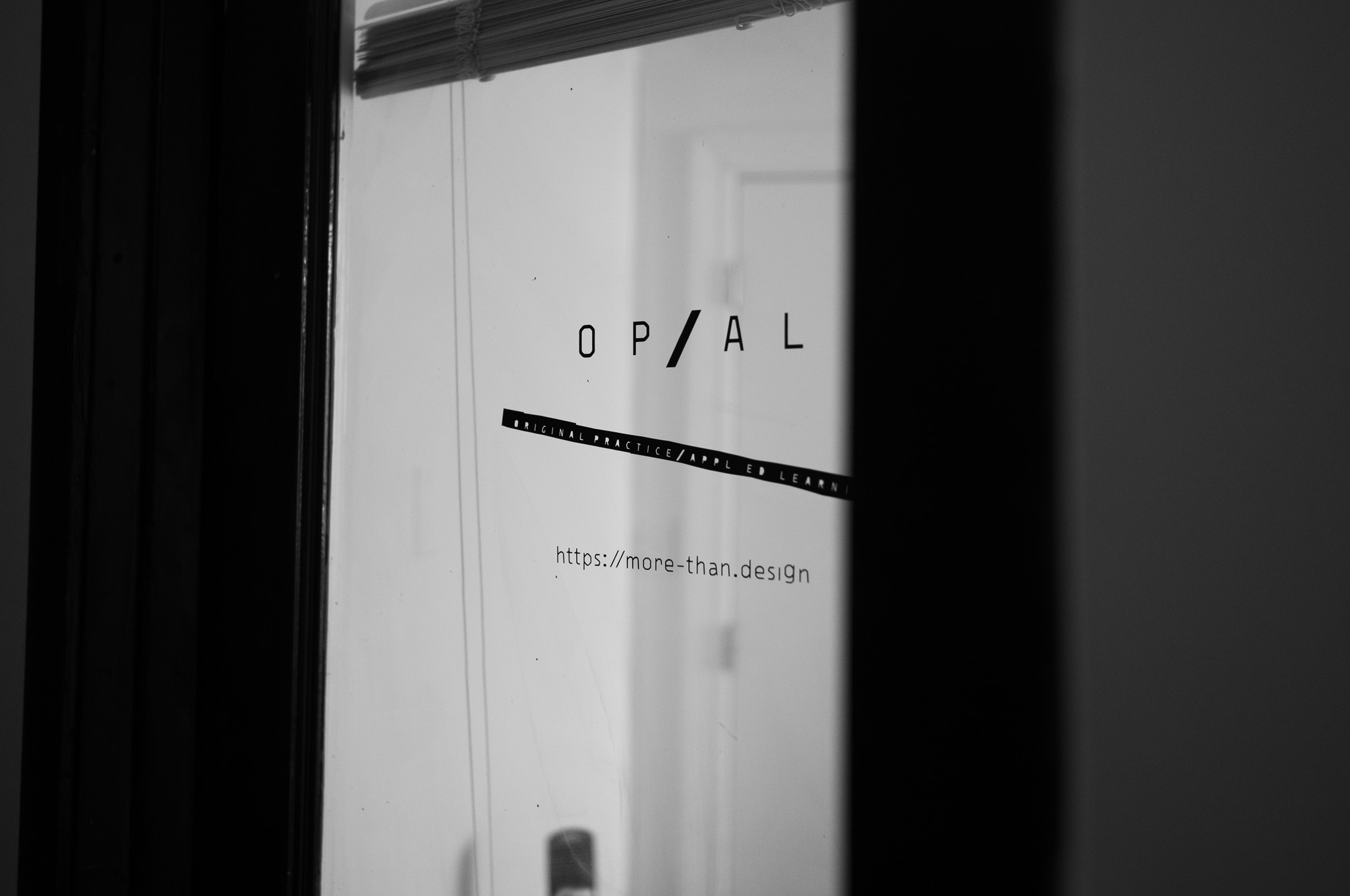 Christian Solorzano — black-and-white photograph of the OP/AL studio glass door etched with the Original Practice / Applied Learning mark and the URL more-than.design