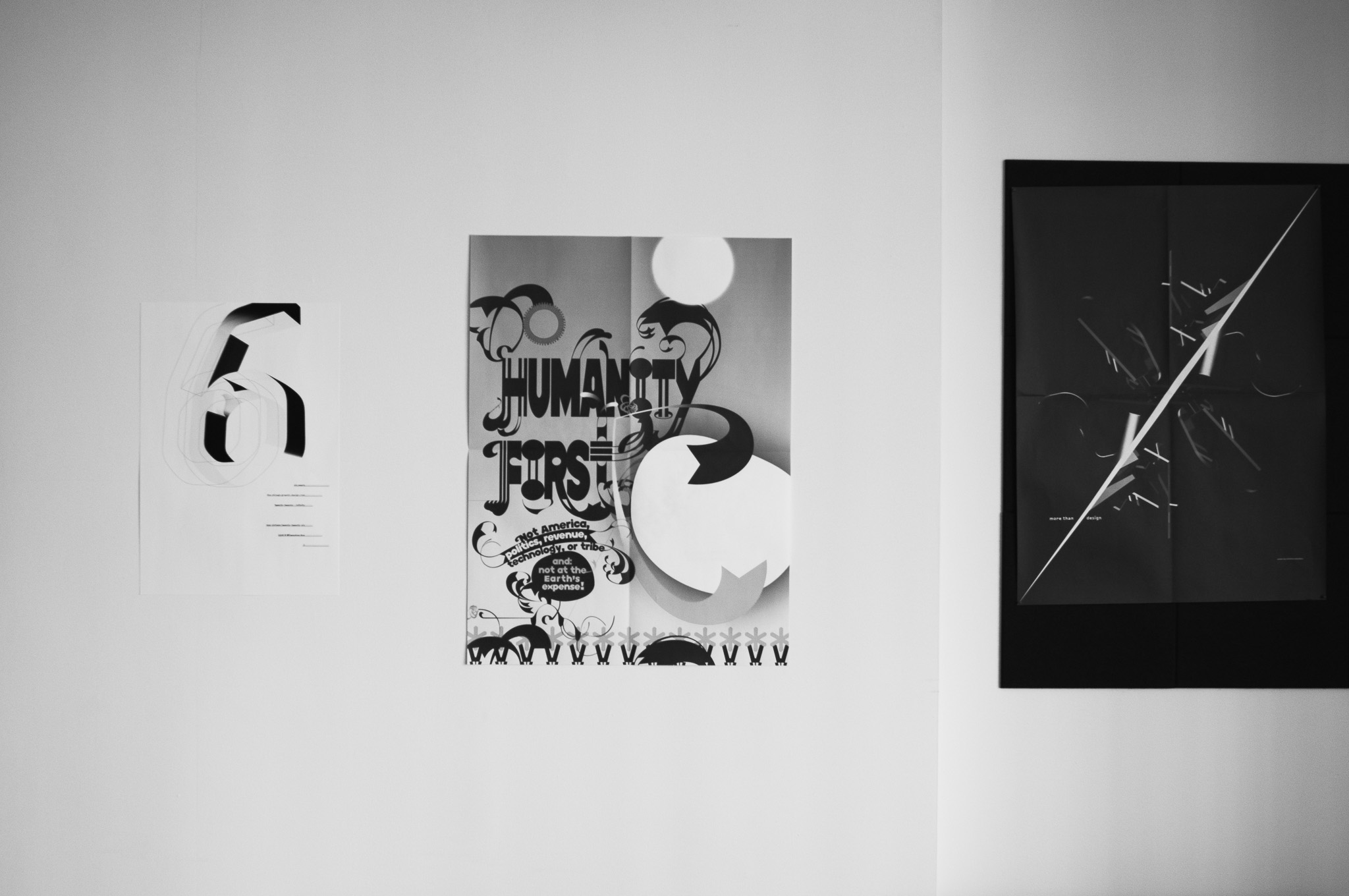 Christian Solorzano — black-and-white photograph of a gallery wall with three posters, including a "Humanity First" typographic print and a dark "more than design" poster