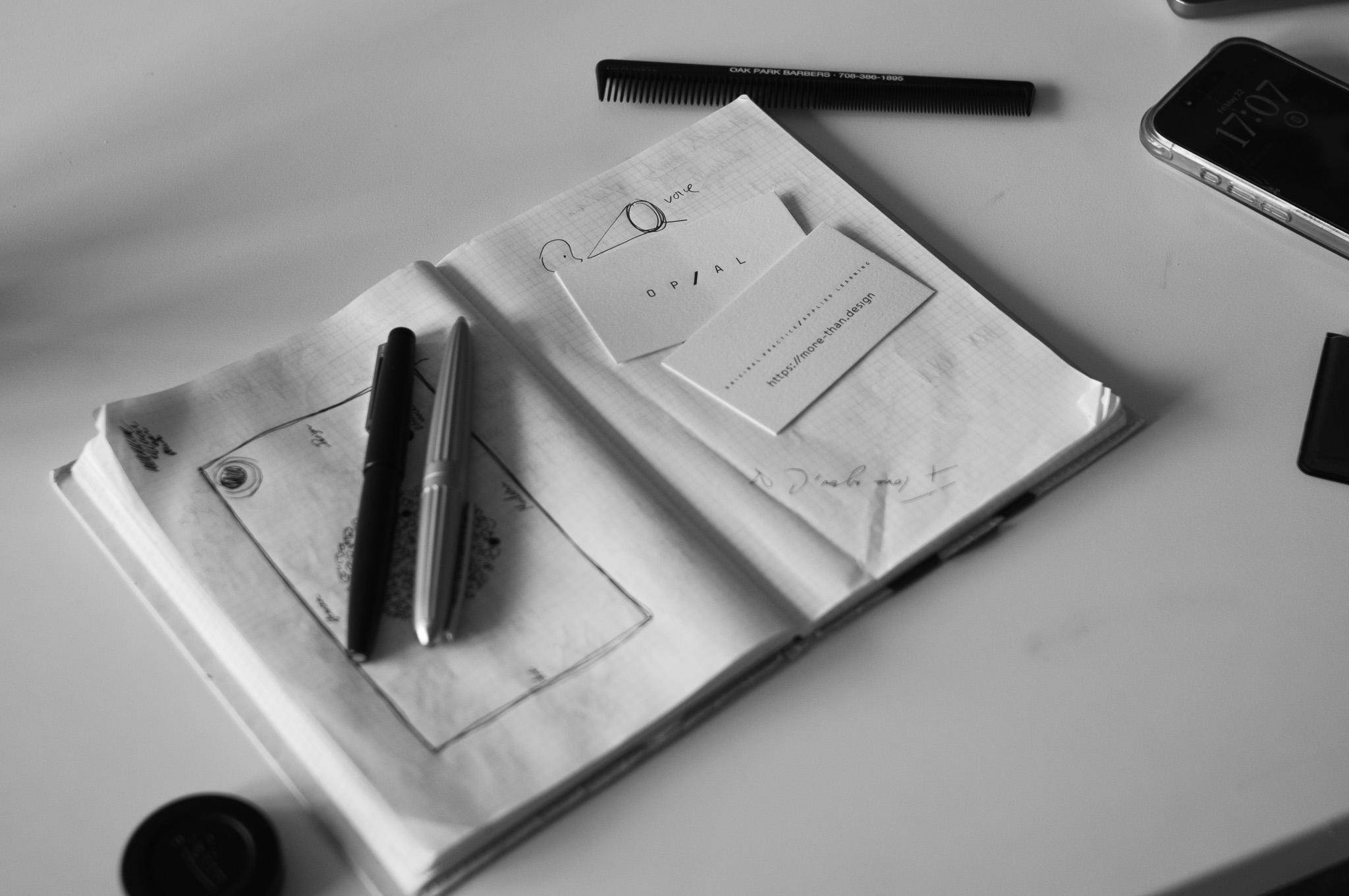 Christian Solorzano — black-and-white desk still life with an open sketchbook, two fountain pens, and OP/AL business cards reading more-than.design