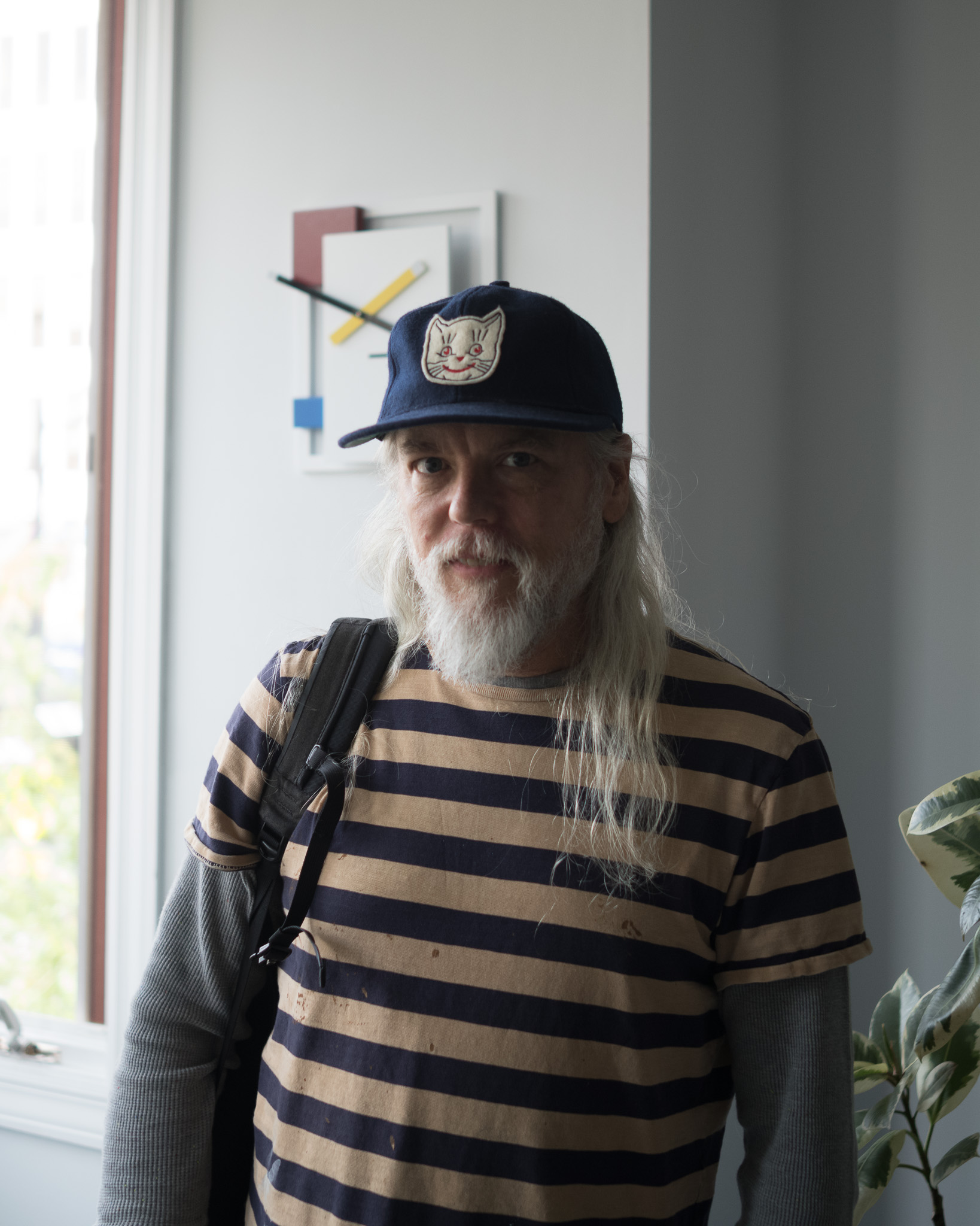 Christian Solorzano — portrait of his friend Brian Morris at OP/AL, wearing a navy cap with a cat patch and a striped shirt, standing by a window