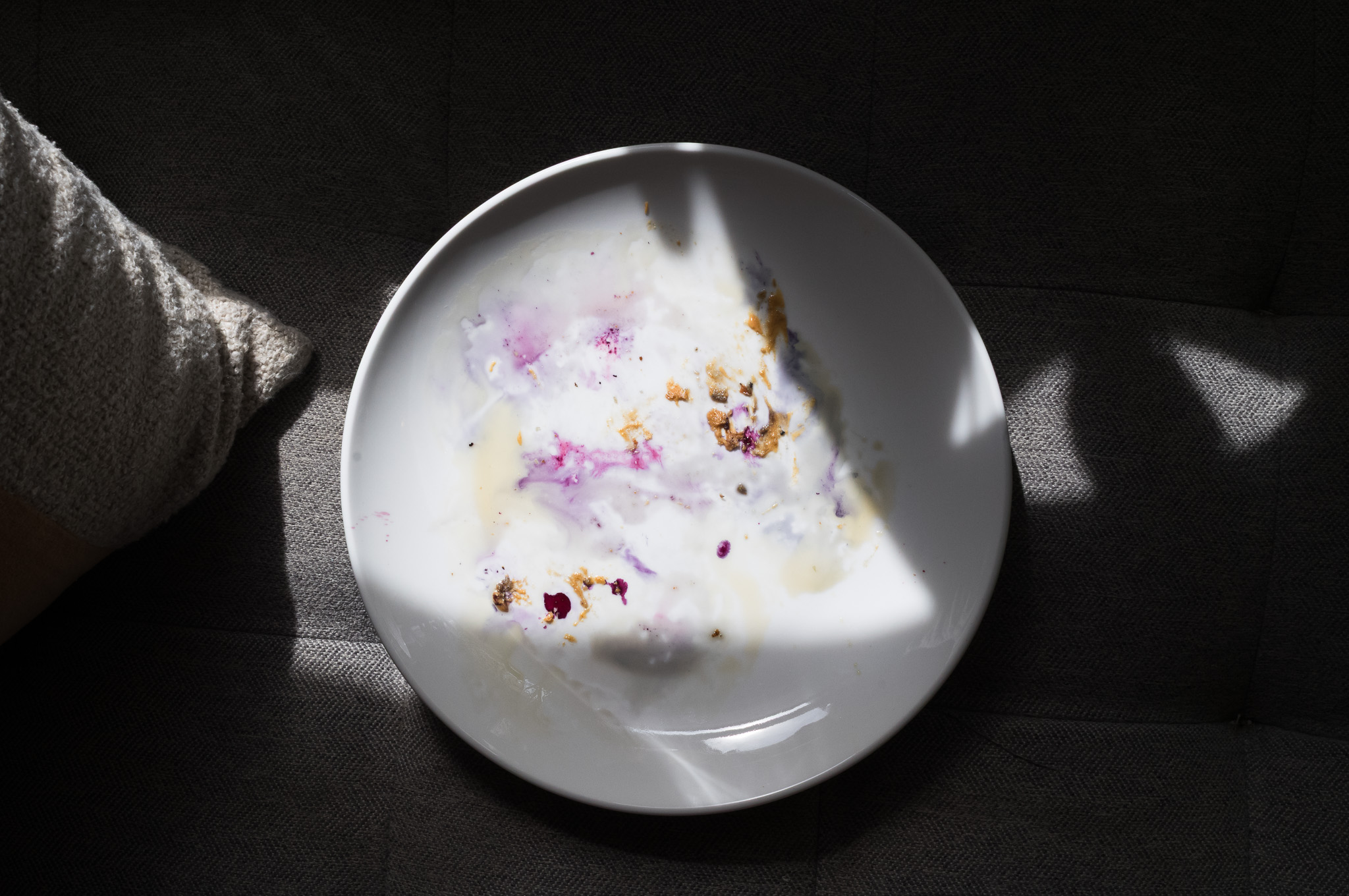 Christian Solorzano — overhead photograph of an empty white bowl with traces of yogurt and berry on a dark grey couch, cut by a shaft of sunlight