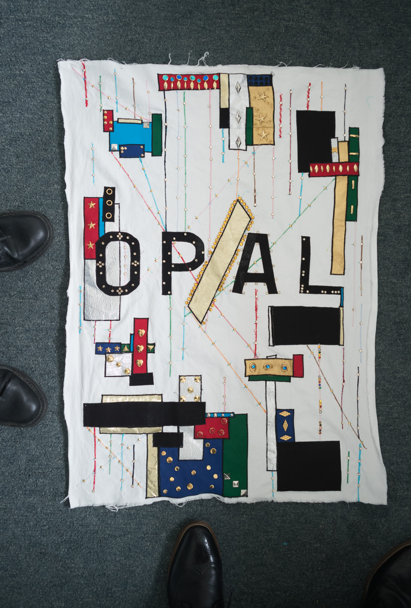Christian Solorzano — photograph of a quilted textile artwork spelling OP/AL in fabric and stitched lines on a grey floor, framed by shoes