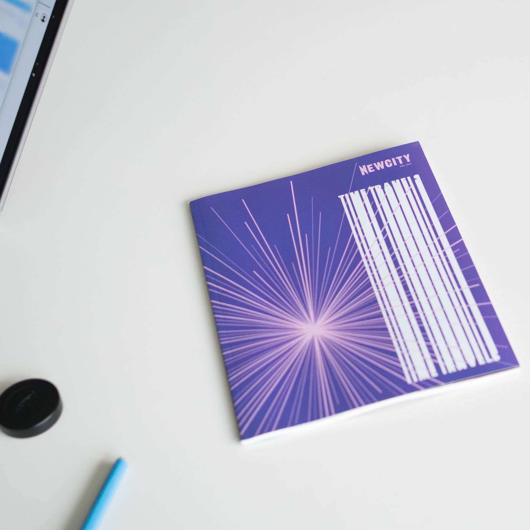 Christian Solorzano — photograph of the NEWCITY "Time Travels" June 2026 cover, a purple starburst with stacked white type, on a desk beside an open laptop