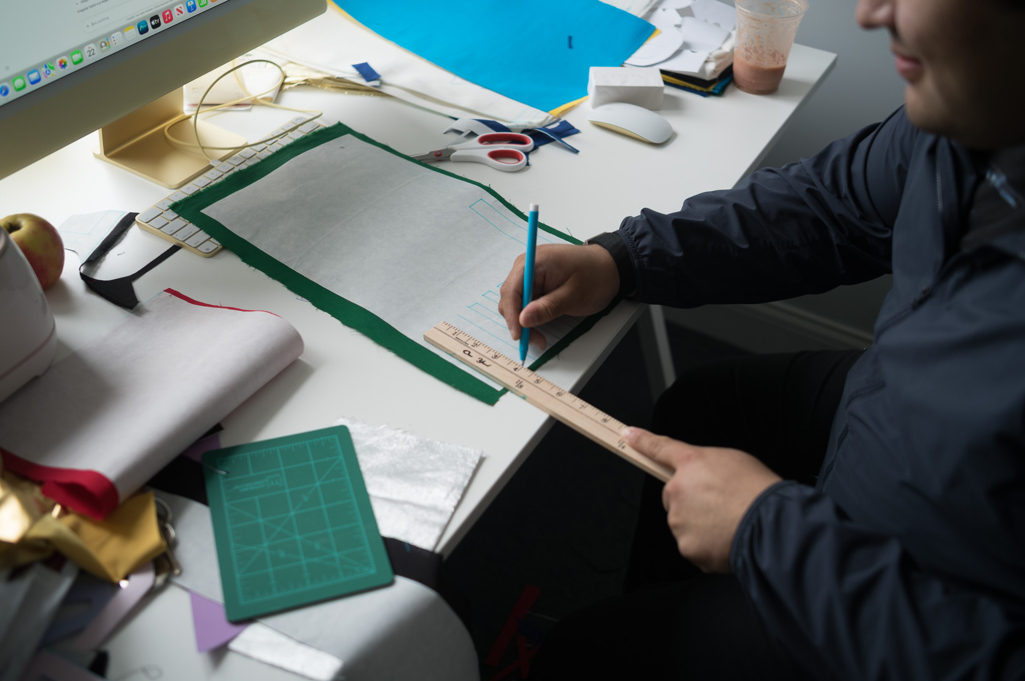Christian Solorzano — photograph of his brother at a desk measuring and cutting felt with scissors and a ruler, with a sewing machine and the OP/AL textile in progress on a green cutting mat