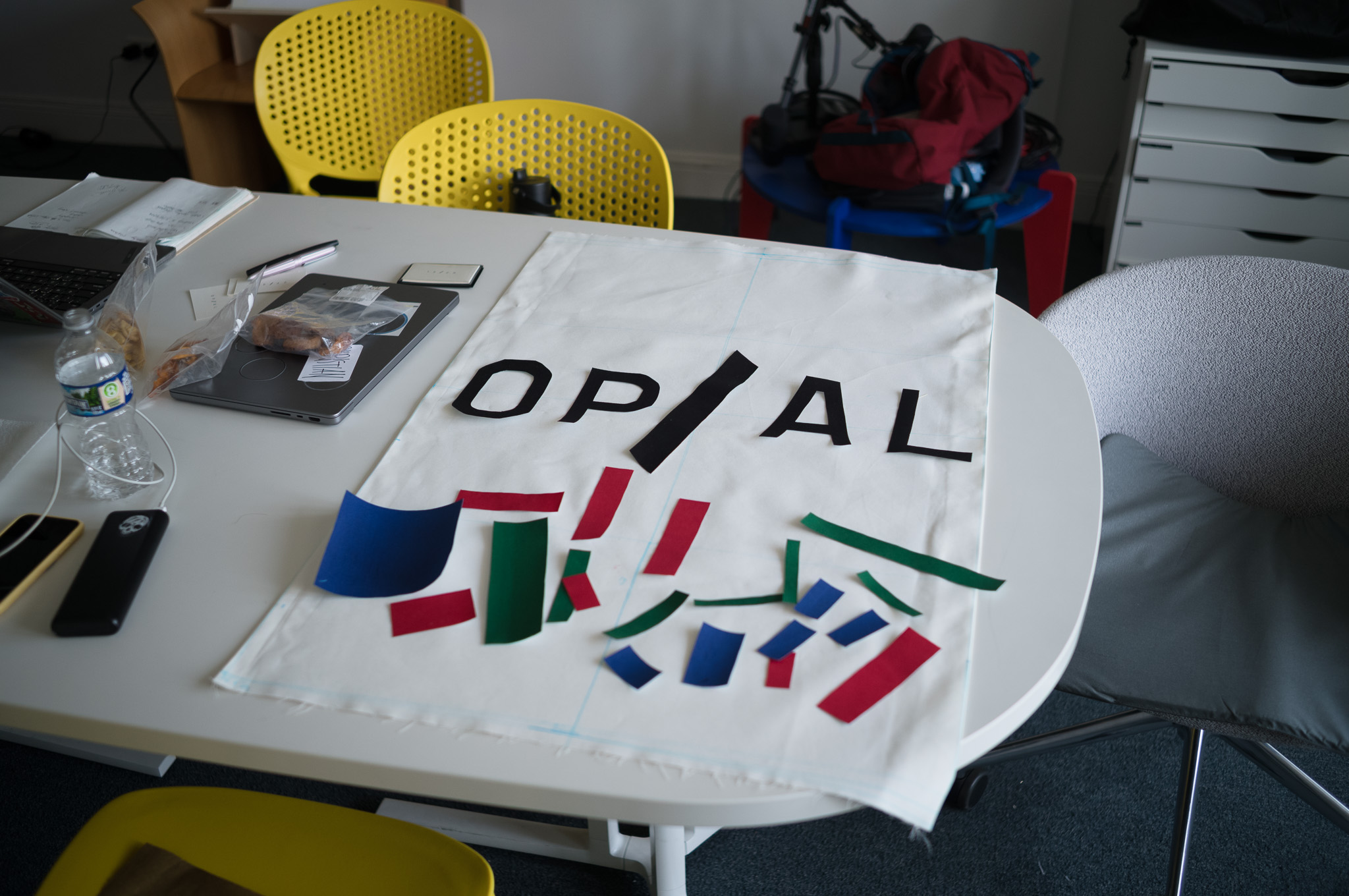 Christian Solorzano — photograph of the OP/AL fabric poster mid-layout on a round white table, with black "OP/AL" type and colored felt pieces being arranged below