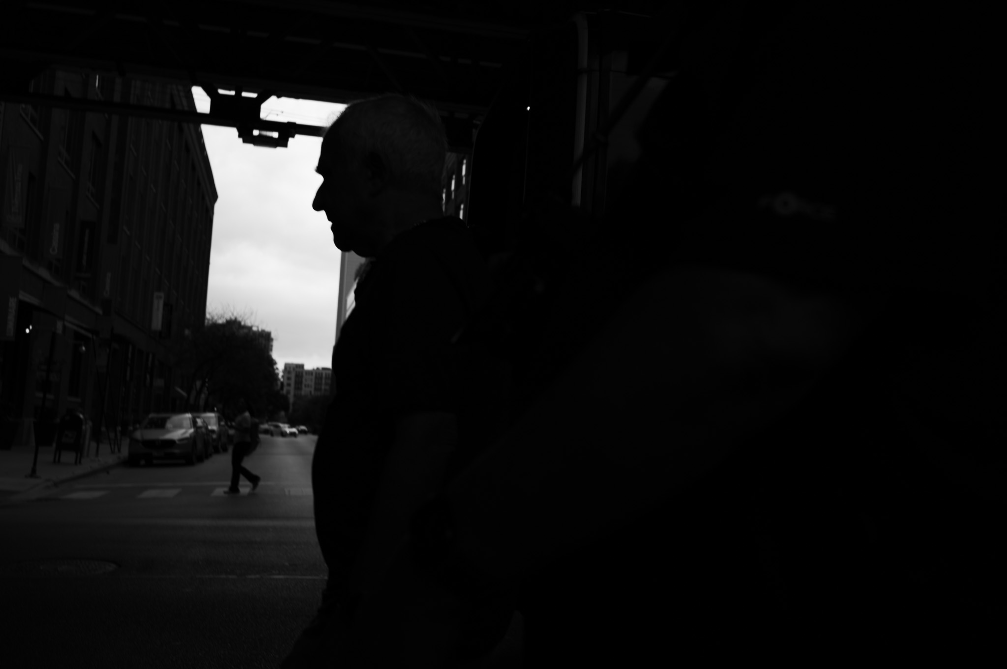 Christian Solorzano — black and white photograph of a man in silhouette walking beneath the elevated tracks, city street receding behind him