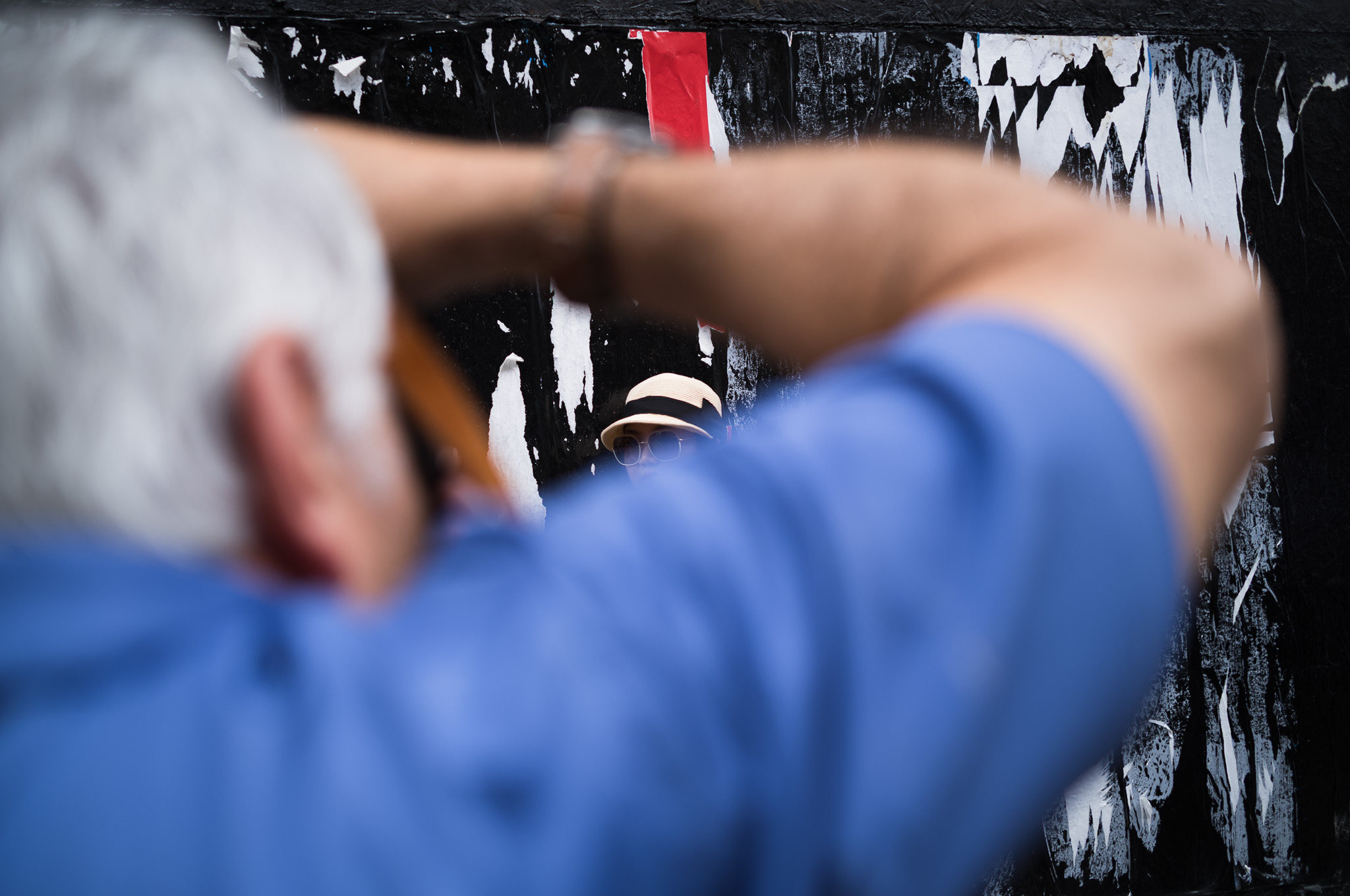 Christian Solorzano — photograph of a man photographing through a gap in a torn poster wall, a figure in a straw hat and sunglasses framed beyond