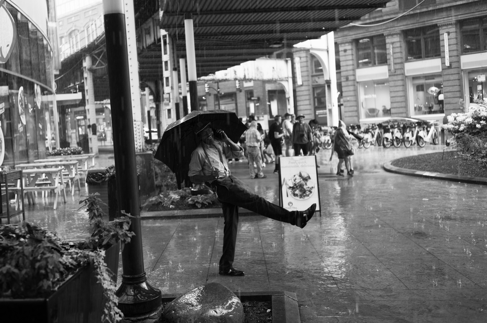 Christian Solorzano — black and white photograph of a man with an umbrella kicking his leg out in the rain beneath the elevated tracks