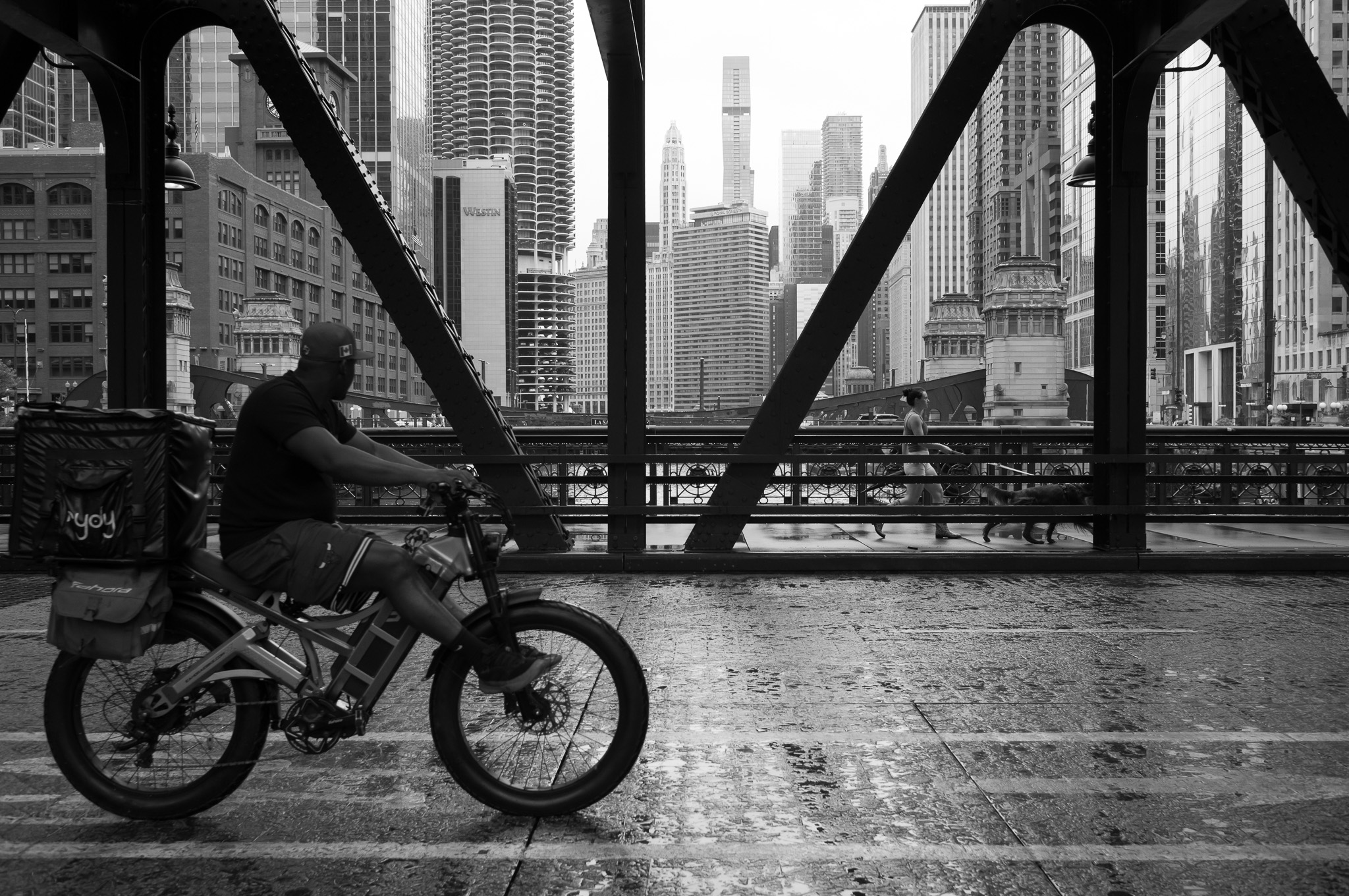 Christian Solorzano — black and white photograph of a delivery cyclist crossing a bridge with the Chicago skyline beyond