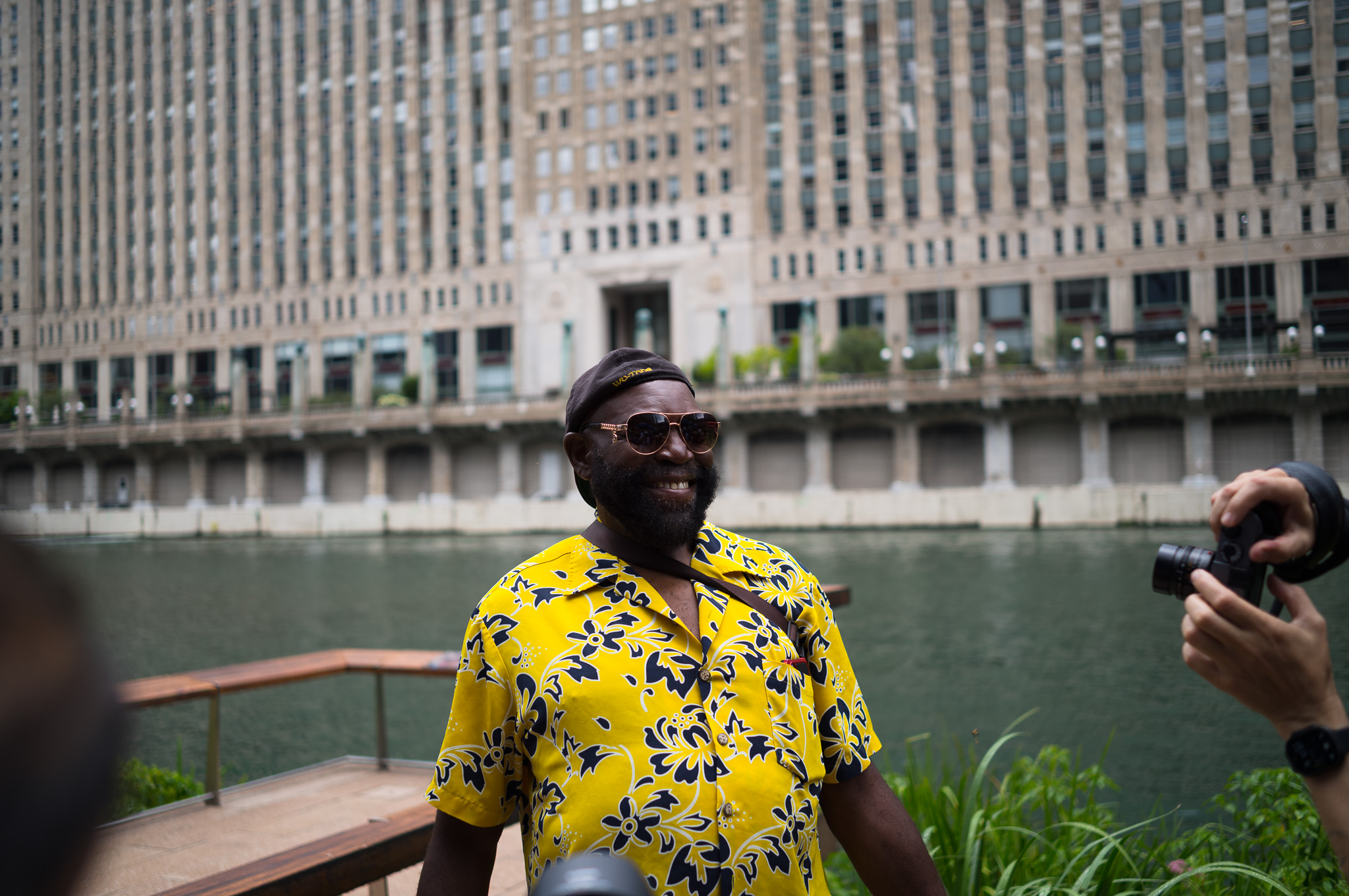 Christian Solorzano — photograph of a man in a yellow floral shirt and sunglasses smiling by the river, the Merchandise Mart behind him