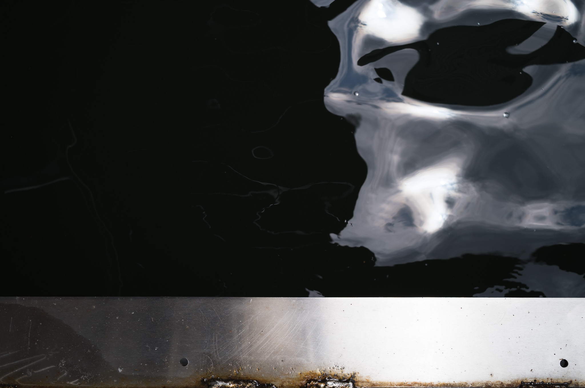 Christian Solorzano — photograph of dark water and reflected light meeting a scratched metal edge