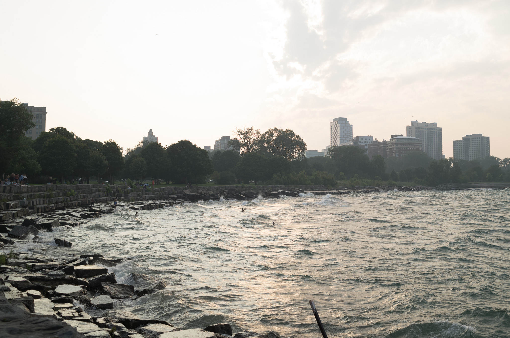 Christian Solorzano — photograph of waves breaking against the rocky lakefront at dusk with the city skyline in haze