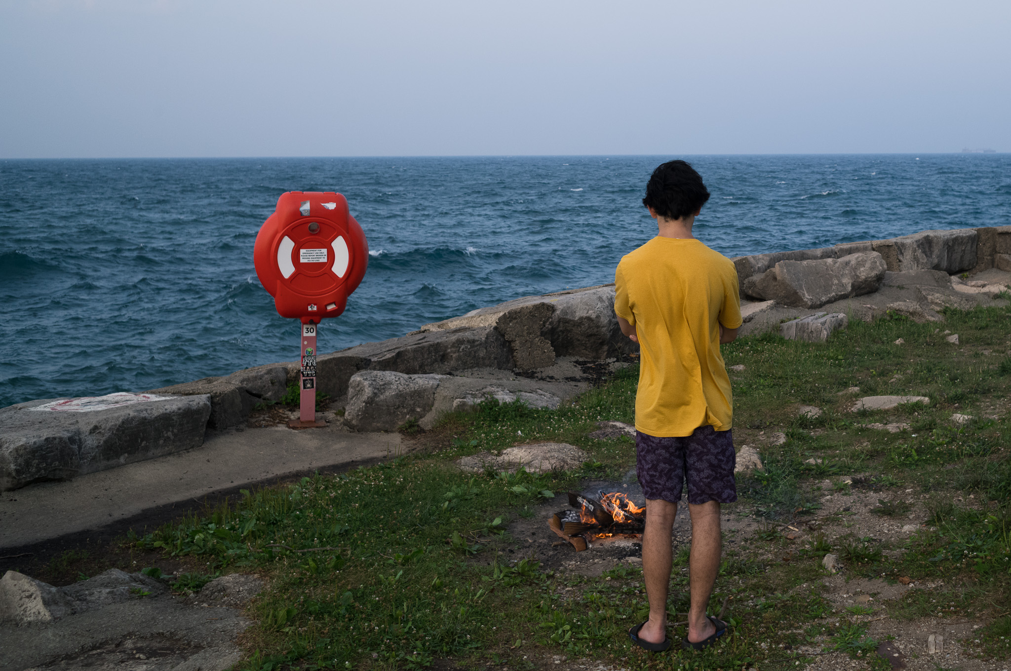 Christian Solorzano — photograph of a person in a yellow shirt standing at the lakefront edge beside a small fire, red life ring station on the shore