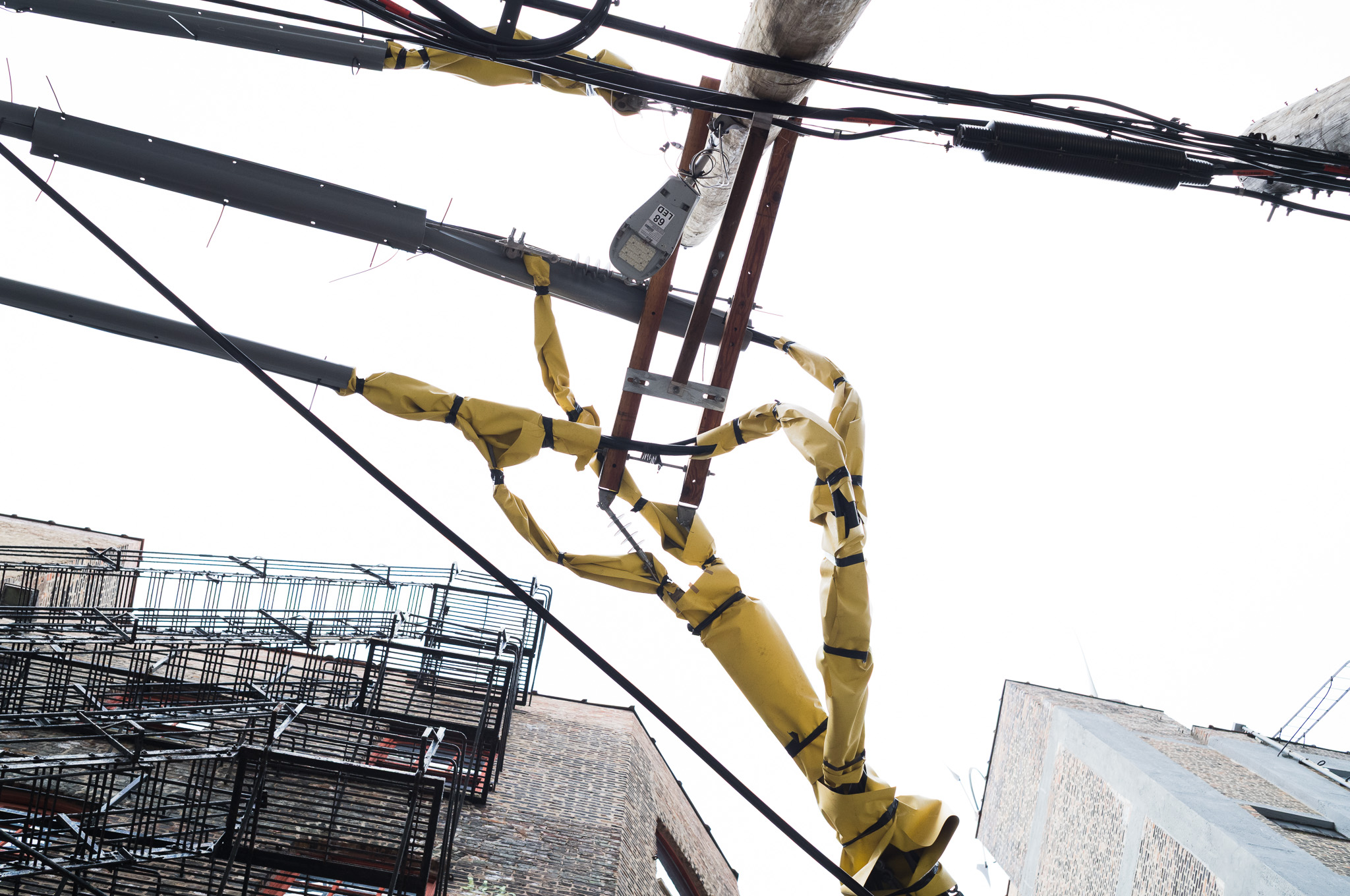 Christian Solorzano — photograph looking up at a utility pole with yellow insulated cable covers twisting against a white sky, fire escapes below