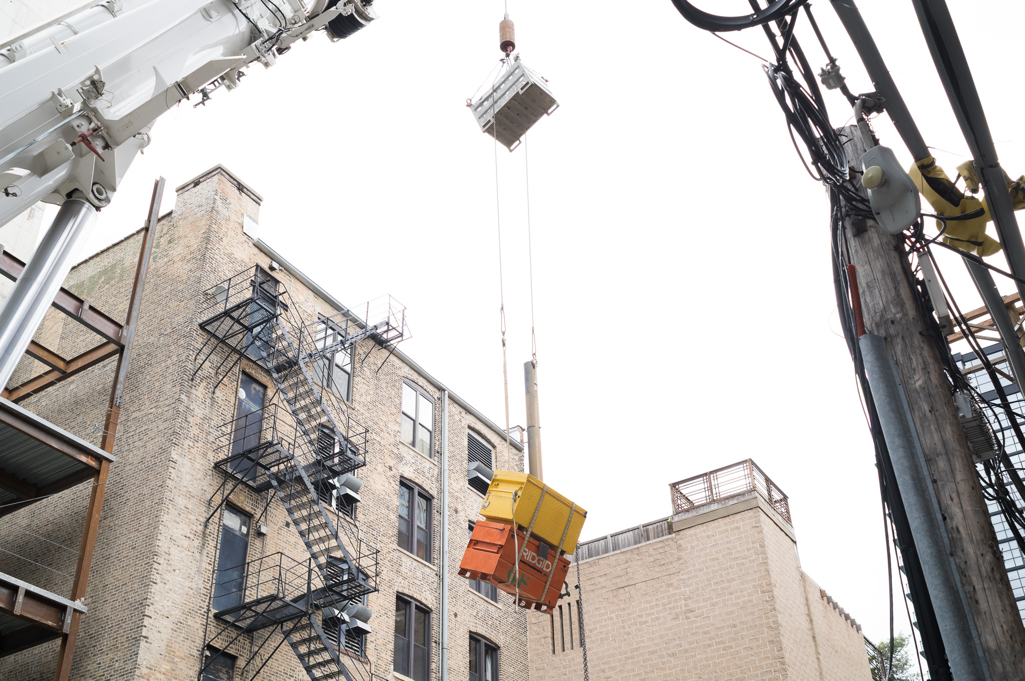 Christian Solorzano — photograph looking up at a crane hoisting a yellow and orange equipment crate above a brick building with fire escapes