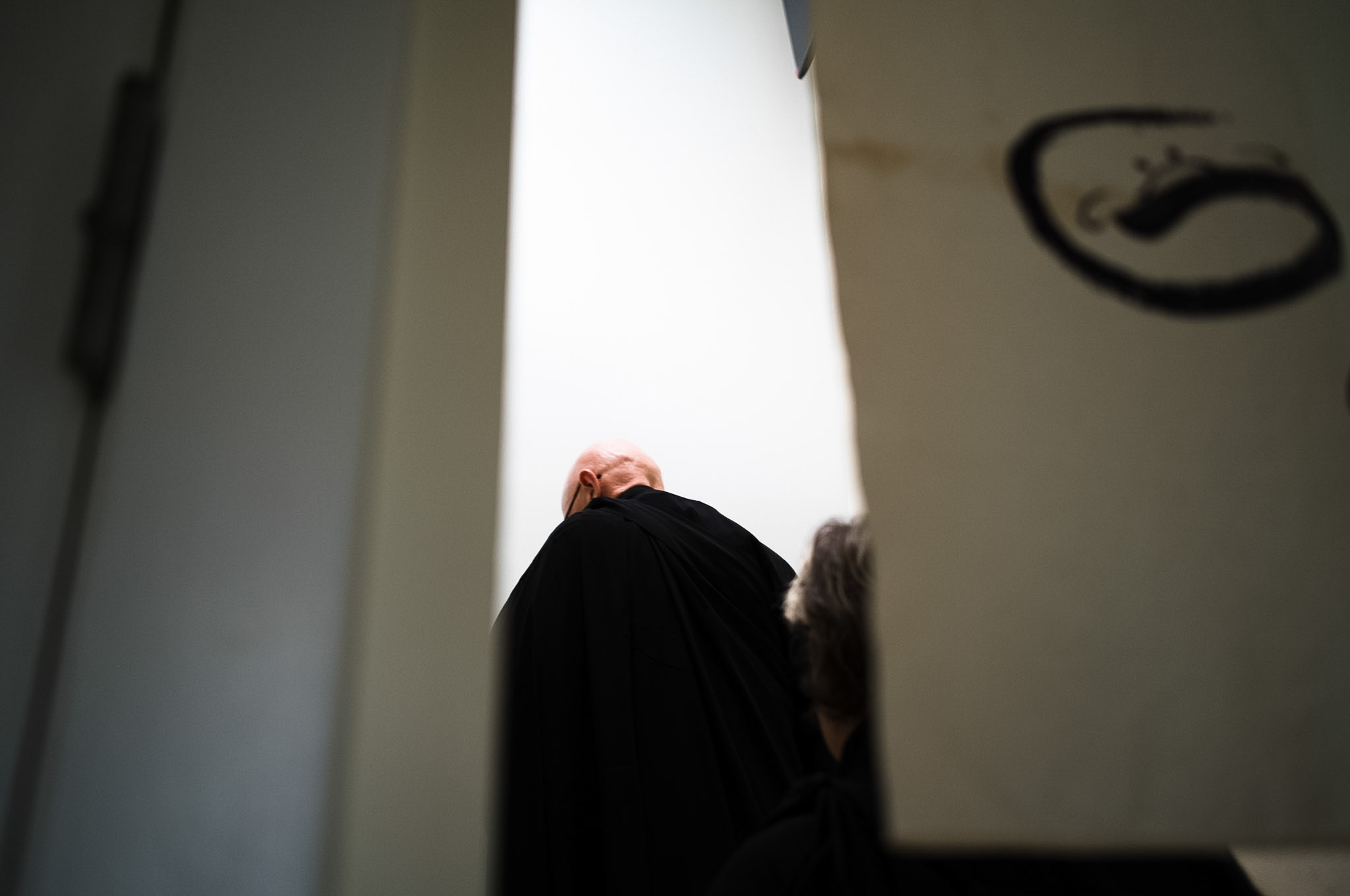 A priest in black robes seen from below, head bowed, against a bright wall with an enso painted on it.