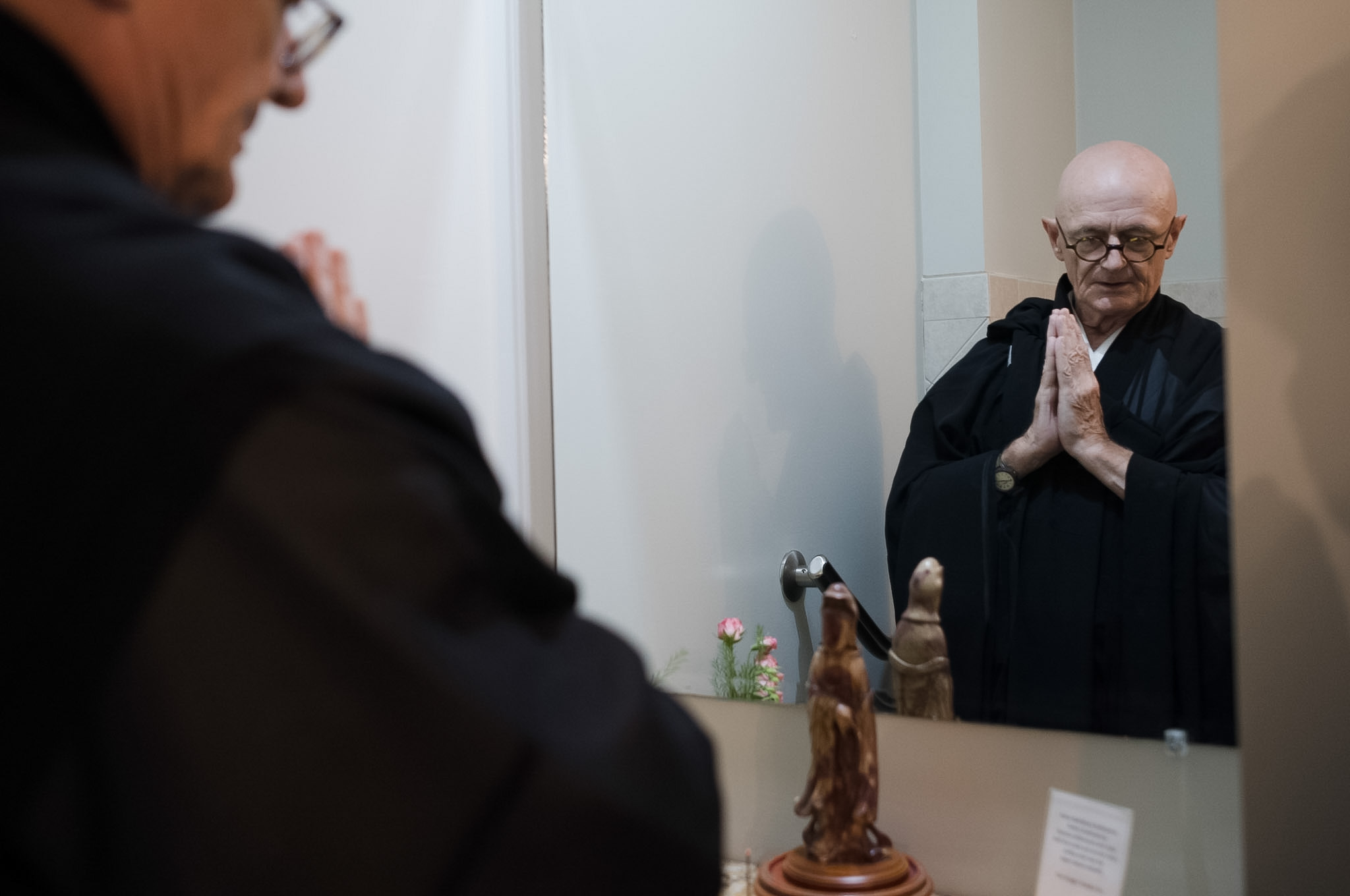 A priest with his hands together in gassho, seen in a mirror over a small altar.