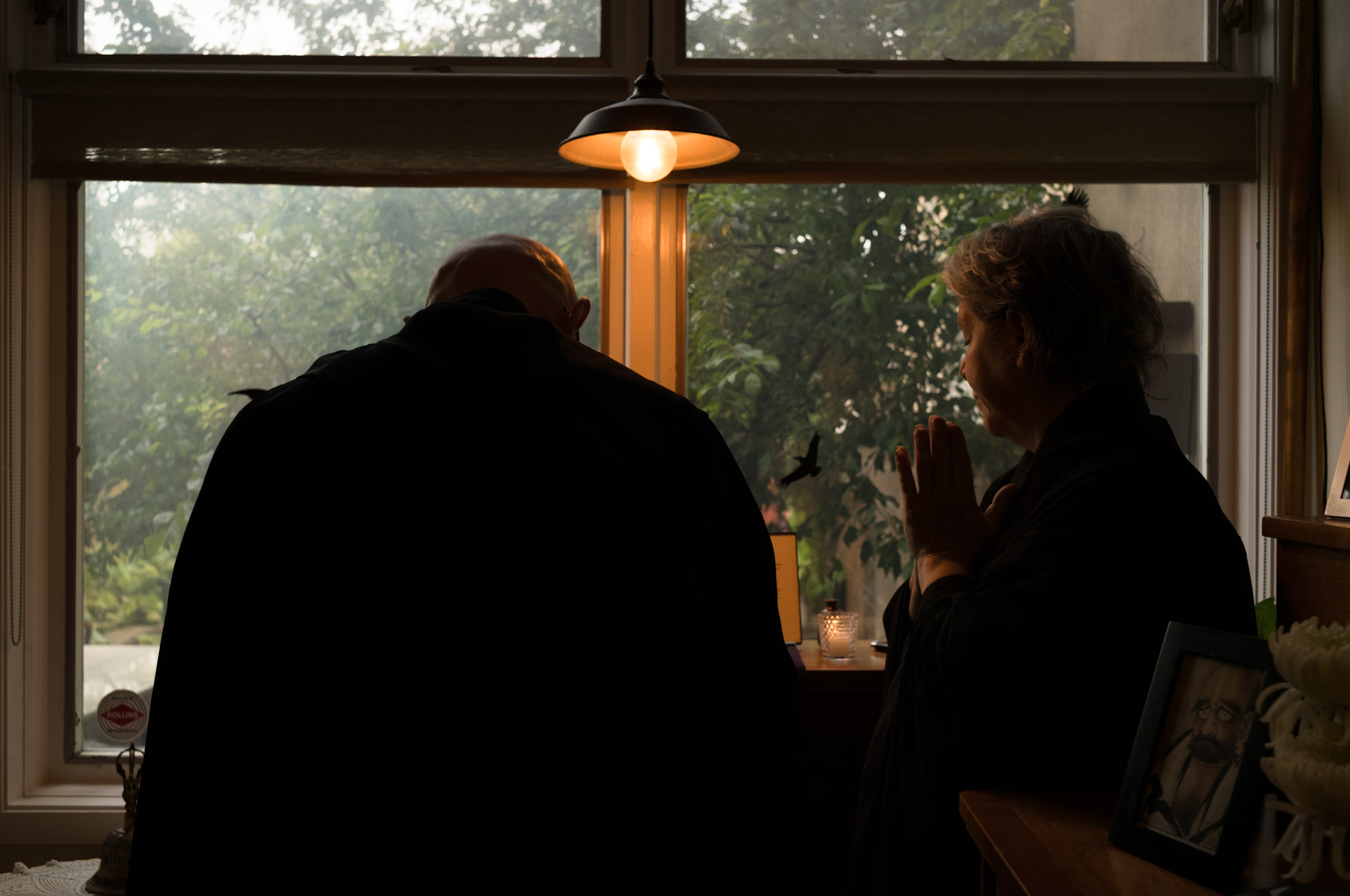 Two people at a window in early light, one bowing, one with hands together.