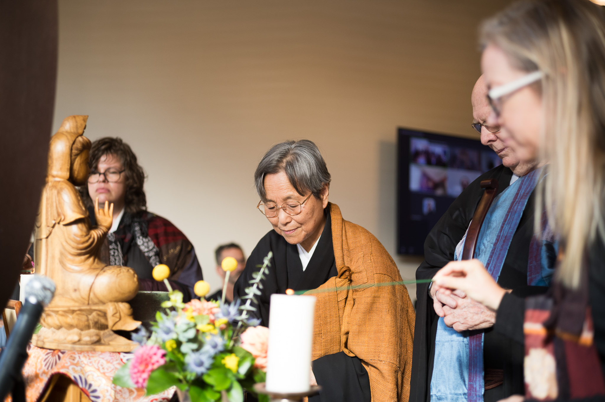 Standing at the altar before a carved wooden bodhisattva, a candle and flowers, hands held together, with others in robes gathered close on either side.