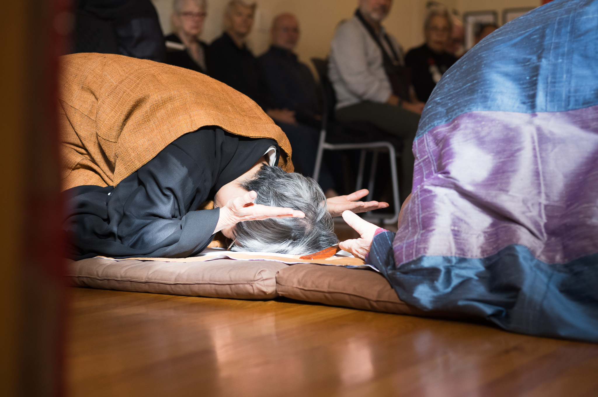 A full prostration on a bowing mat, forehead to the floor and palms turned up, seen low across the wood floor with the seated assembly behind.