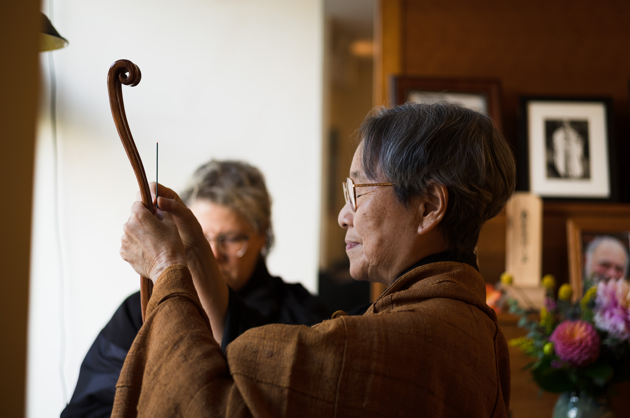 Seen in profile in an ochre kesa, holding a curved wooden nyoi upright at arm's length, with a second person behind and framed portraits and flowers on the altar beyond.