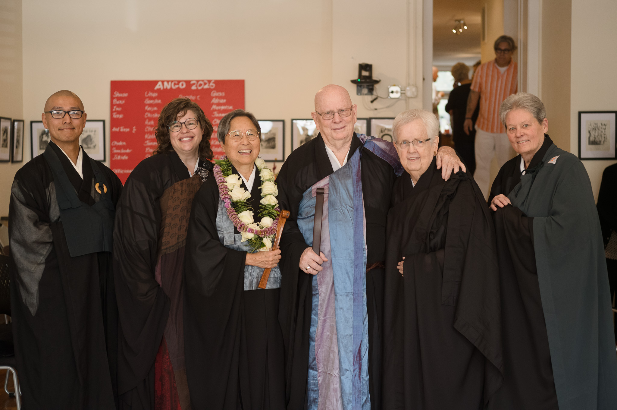 Six people in robes standing together for a portrait after the ceremony, one wearing a white flower lei, the Ango 2026 board on the wall behind.