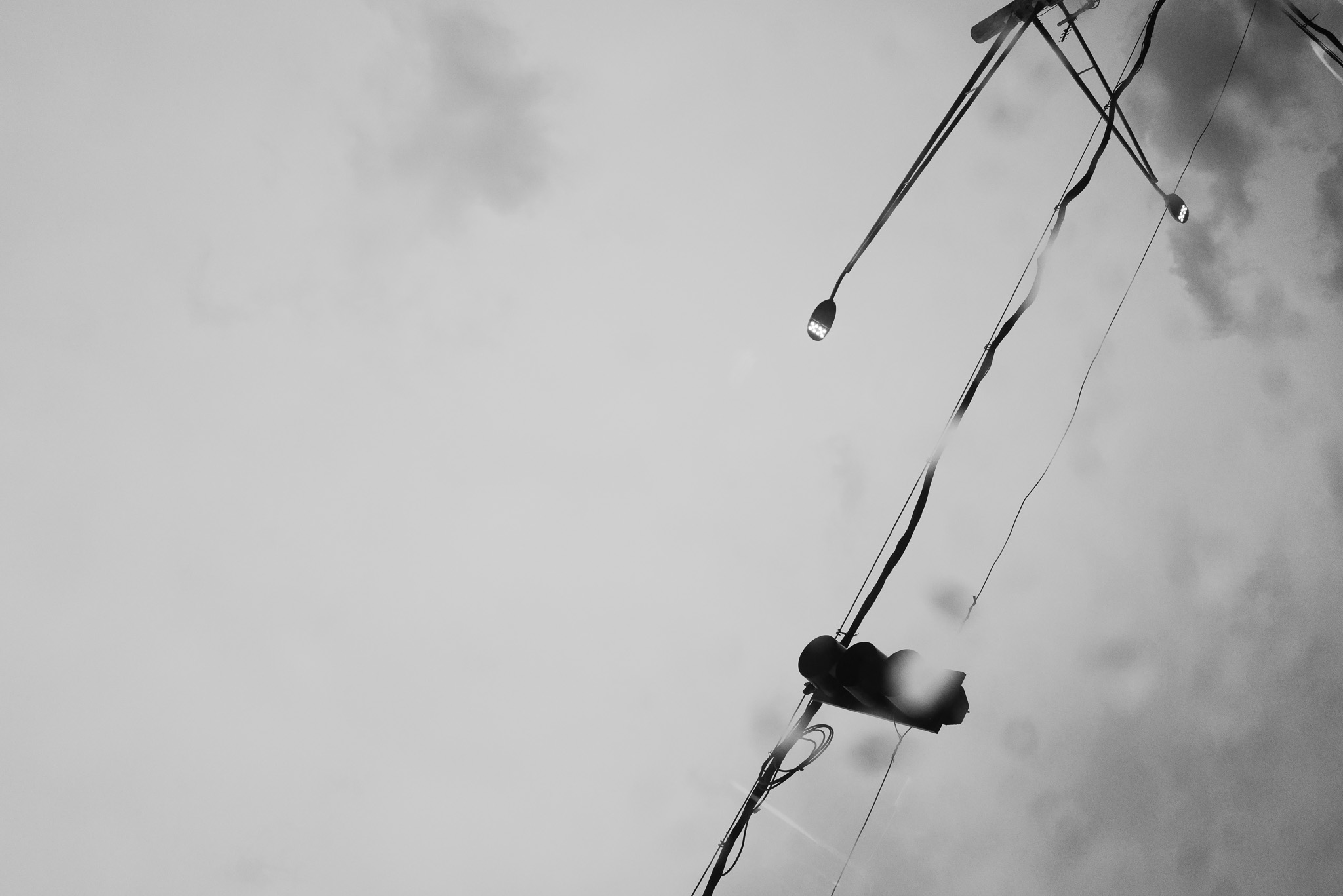 Christian Solorzano — black-and-white photograph looking up at silhouetted streetlights and tangled overhead wires against an overcast sky