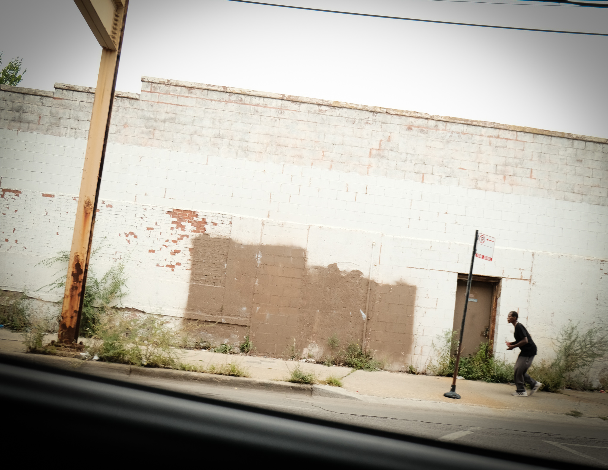Christian Solorzano — street photograph of a man walking past a patched white brick warehouse wall, shot from a passing car