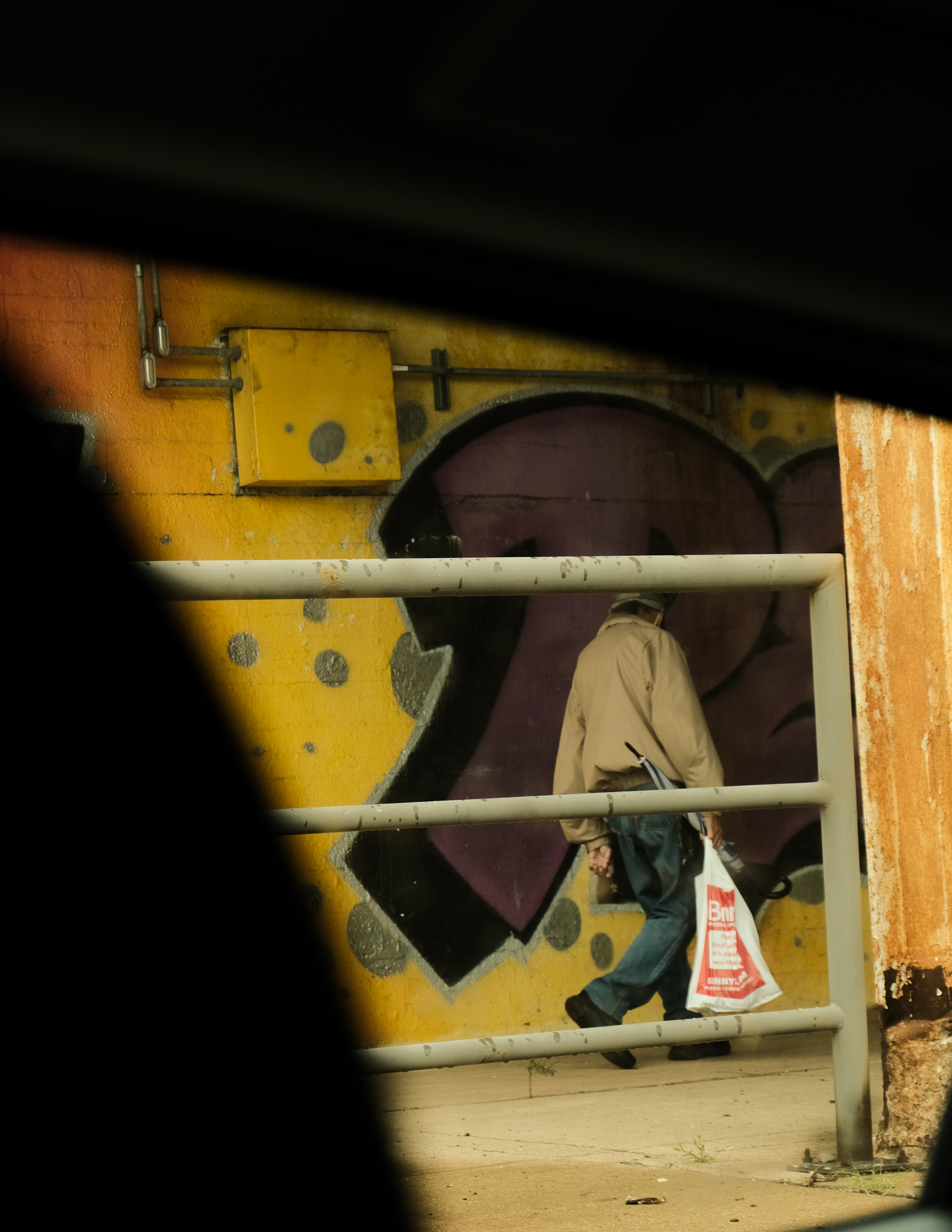 Christian Solorzano — street photograph of an older man with a plastic bag walking past a yellow graffiti wall beneath an overpass