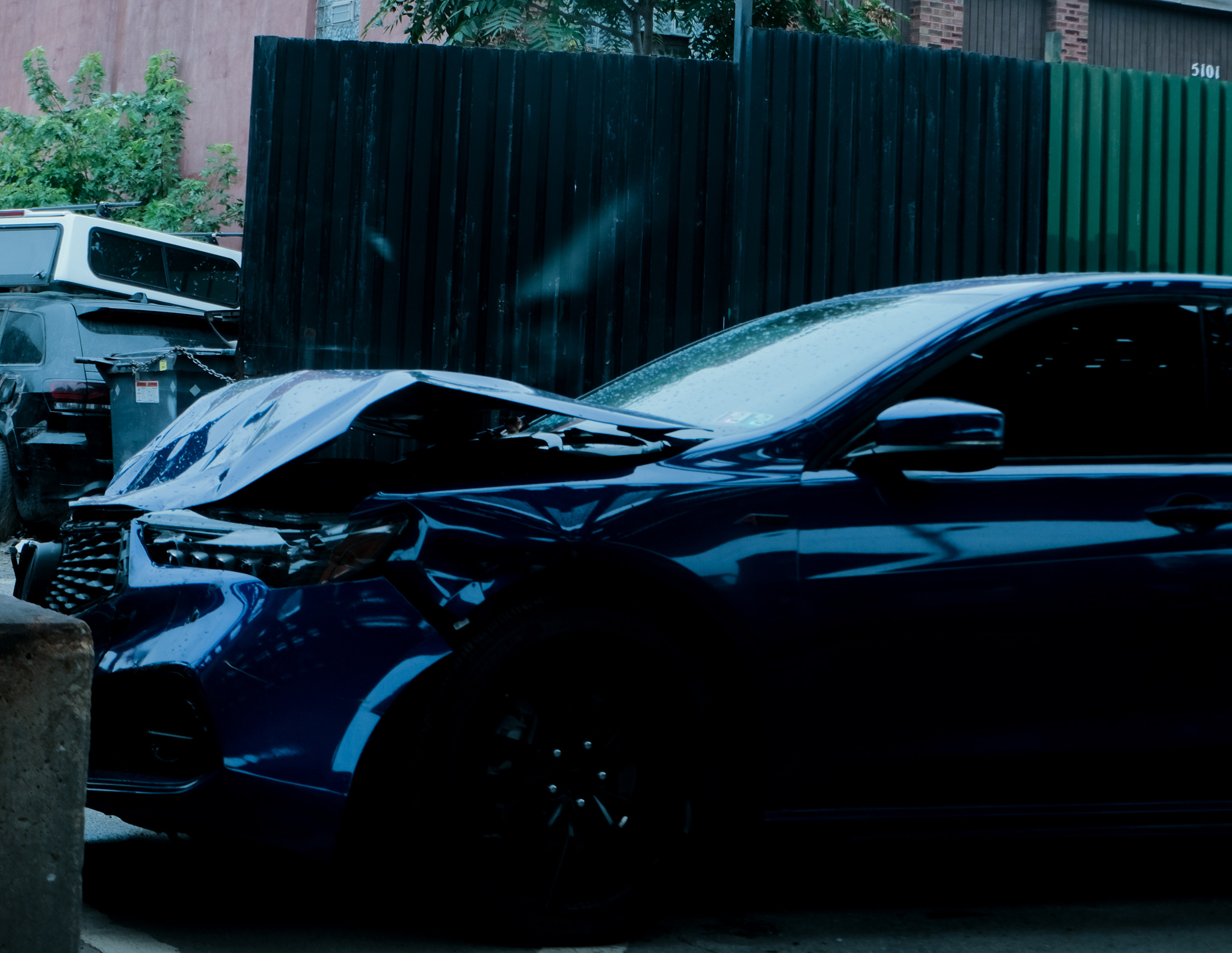 Christian Solorzano — photograph of a dark blue sedan with a crumpled hood parked beside a black fence in an alley
