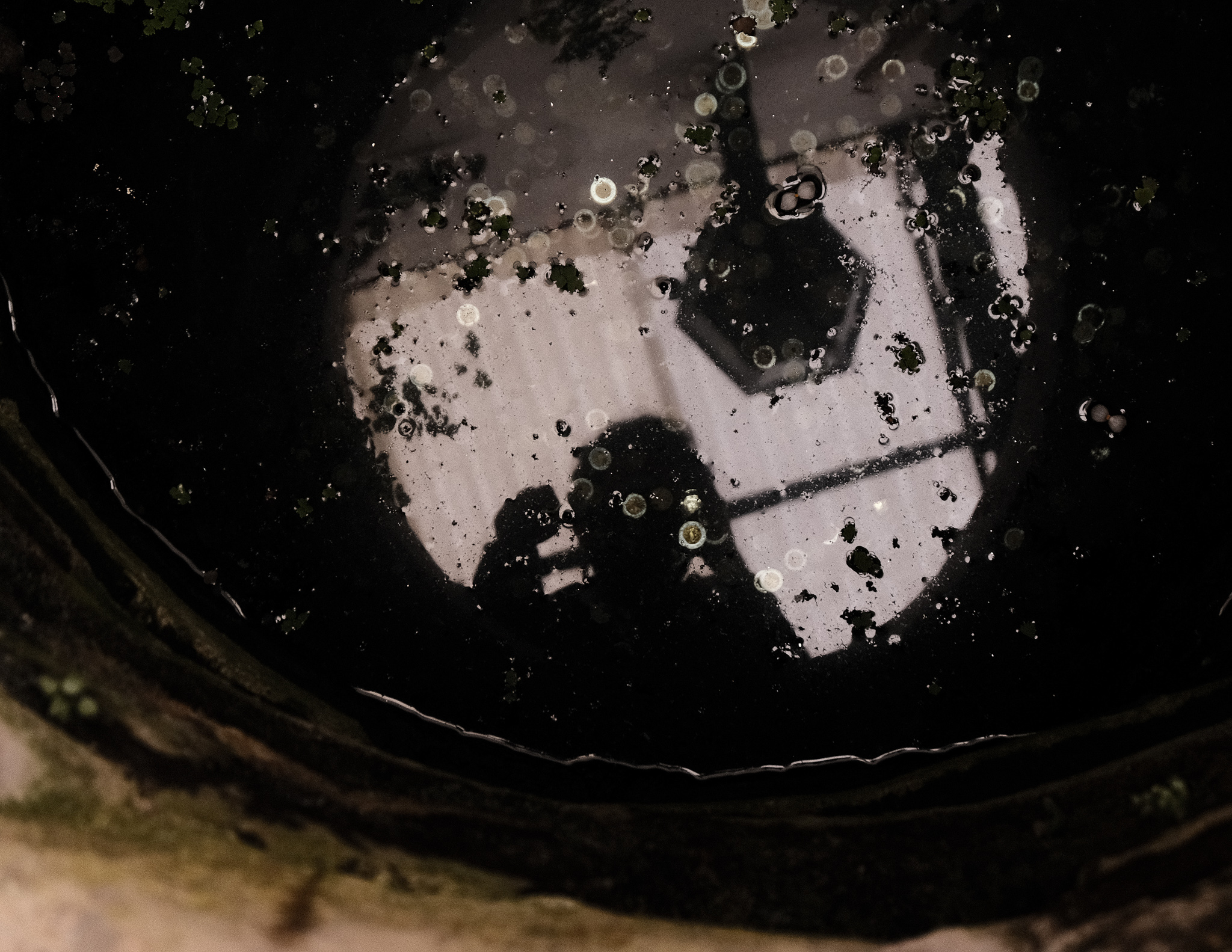 Christian Solorzano — self-portrait reflected in dark water as the photographer crouches with a camera
