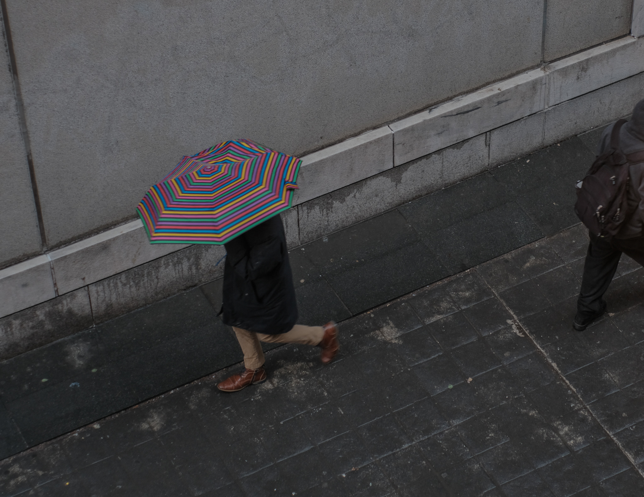 Christian Solorzano — overhead street photograph of a person with a rainbow-striped umbrella on a wet sidewalk