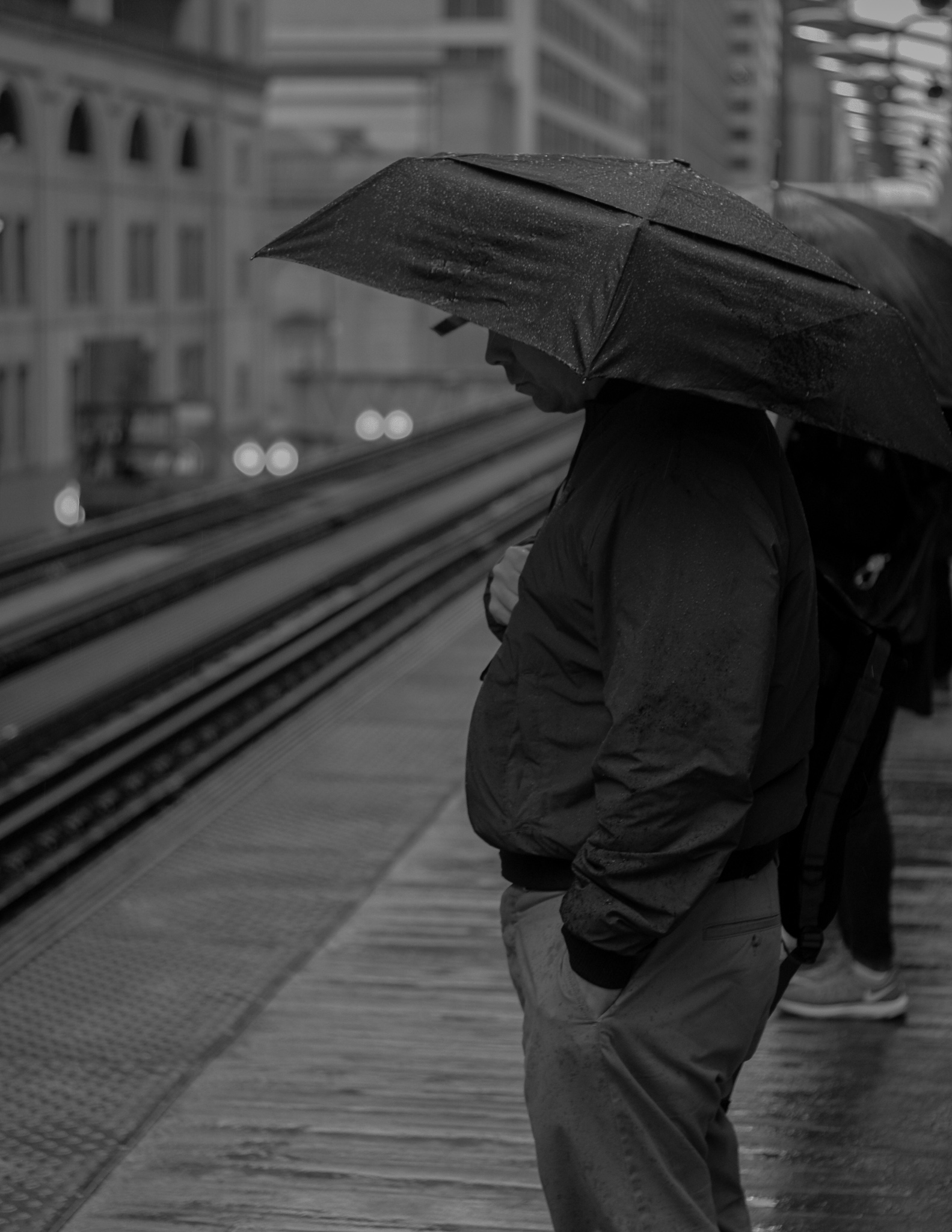 Christian Solorzano — black-and-white photograph of a man under an umbrella waiting on a rainy Chicago L platform